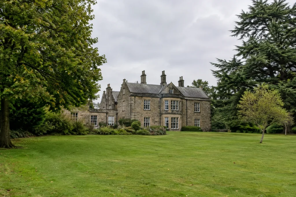 A generic stone country house seen across a lawn, with mature trees and an overcast sky, without signs or identifiable people.