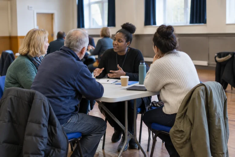 Anonymous adults chatting beside a folding table inside a community hall, with coats on chairs and no visible signs