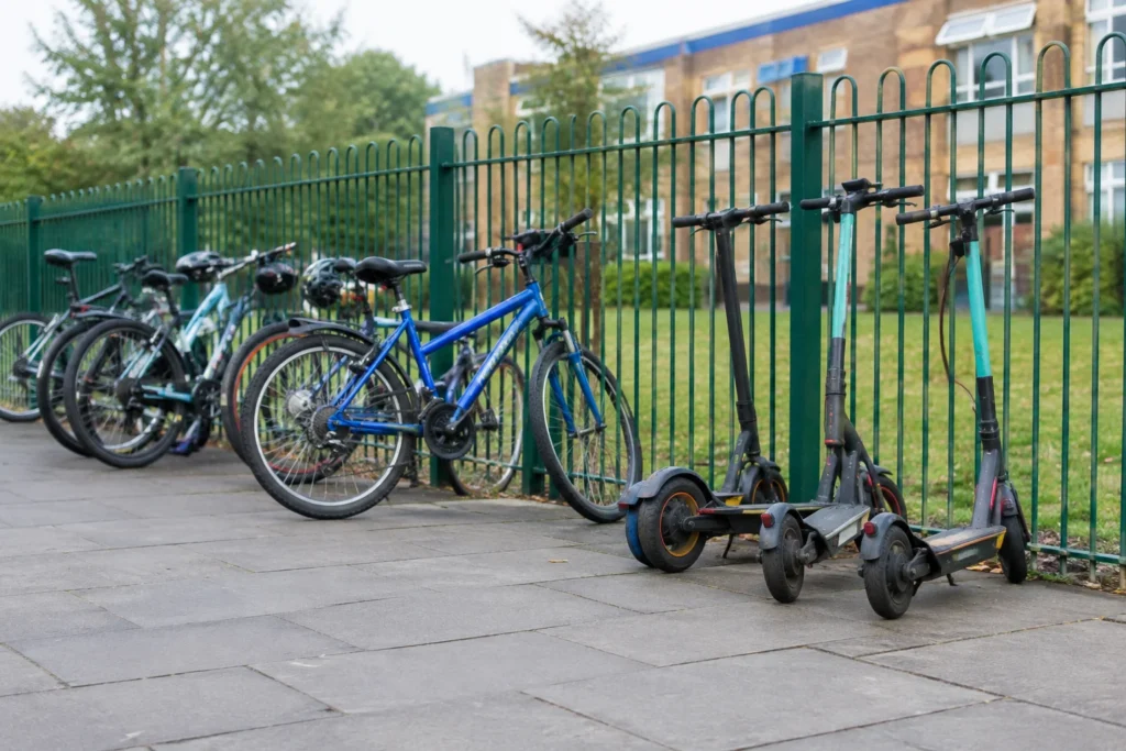 Bicycles and scooters parked beside a plain school-style fence, with a pavement in the foreground and no readable signs.