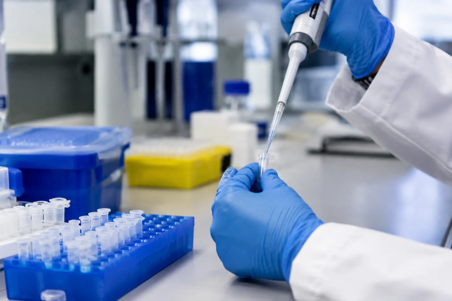 A close view of anonymous gloved hands using a pipette beside clear sample tubes on a laboratory bench.