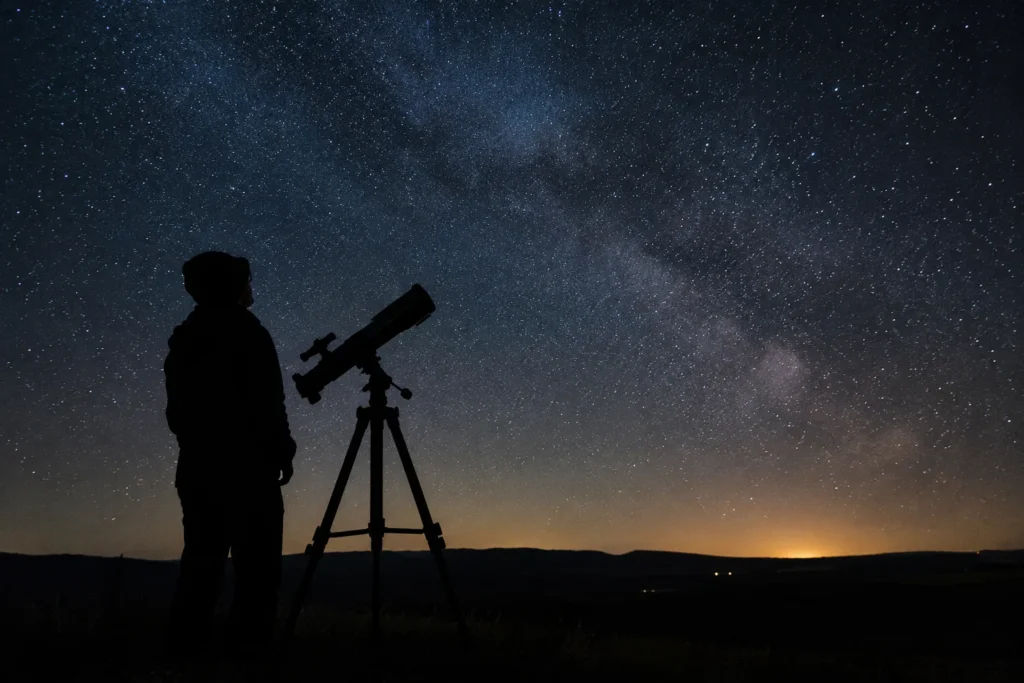 An anonymous observer beside a telescope silhouetted against a rural night horizon.