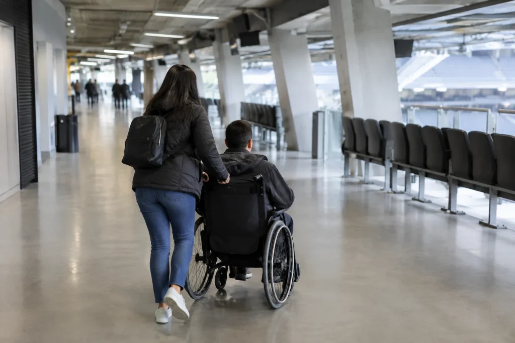 An anonymous wheelchair user and companion moving along a broad step-free concourse with plain seating nearby