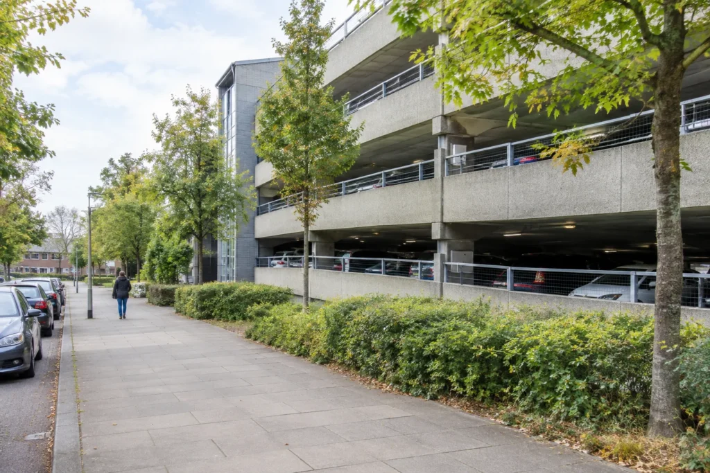 A generic multi-storey car park beside a pedestrian pavement, with parked cars and trees in soft daylight